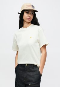 Persona joven con cabello largo y oscuro que lleva una gorra beige, camiseta blanca hueso con un pequeño logo y pantalones vaqueros oscuros, con las manos en los bolsillos, mirando hacia la derecha.