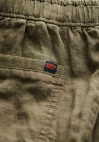 Tissu en lin vert avec une texture froissée et une taille élastique. Présente une petite étiquette "Superdry" rouge et bleue sur la couture latérale.