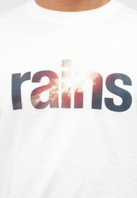 Κοντινό πλάνο λευκού πουκάμισου με τη λέξη «rains» τυπωμένη με μεγάλα γραφιστικά γράμματα που απεικονίζουν έναν συννεφιασμένο ουρανό με ηλιαχτίδες.