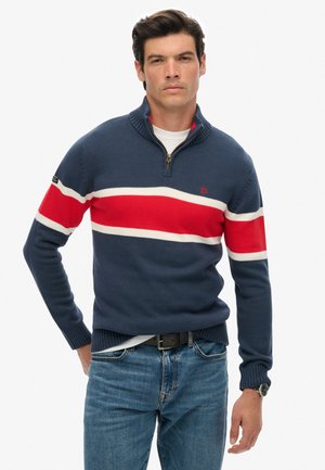Granatowy sweter z dzianiny z kołnierzykiem na zamek. Ozdobiony poziomymi paskami w kolorze czerwonym i białym. Noszony na białej koszulce, w połączeniu z niebieskimi dżinsami.
