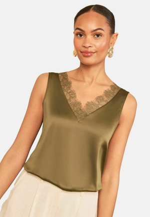 Femeie purtând un top verde din satin fără mâneci cu decolteu în V din dantelă și pantaloni bej, accesorizată cu cercei mari aurii.