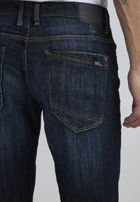 Tamno plave traperice s žutim šavovima, koje imaju dva stražnja dzepa i malu oznaku na desnom dzepu. Čvrst materijal i jednostavan dizajn.