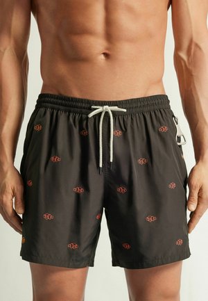 Mannelijke torso met zwarte zwembroek met kleine oranje clownvis patronen en een witte koordsluiting in de taille.