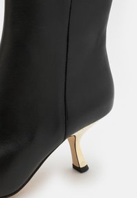 Botte de cheville en cuir noir avec une texture lisse, un talon doré effilé et des coutures discrètes. Design simple avec un bout arrondi.