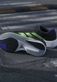 Μαύρα και πράσινα παπούτσια τρεξίματος Adidas με λευκές σόλες, τοποθετημένα σε άσφαλτο με θολή διάβαση πεζών στο φόντο.