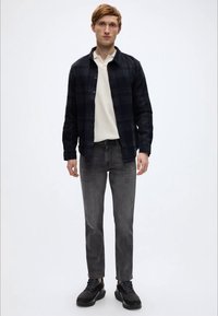 Μαύρο καρό πουκάμισο με κουμπιά πάνω από μπλουζάκι πόλο σε κρεμ απόχρωση, συνδυασμένο με γκρι τζιν και μαύρα αθλητικά παπούτσια. Το μοντέλο στέκεται μπροστά σε λευκό απλό φόντο.