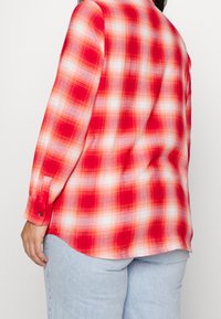 Osoba ubrana w czerwoną i białą koszulę w kratę z długim rękawem, zestawioną z jasnoniebieskimi dżinsami, stoi tyłem na tle jednolitego tła.