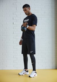 Man in a black sports t-shirt and black shorts holds a kettlebell. He wears black athletic leggings with white accents and white sneakers.