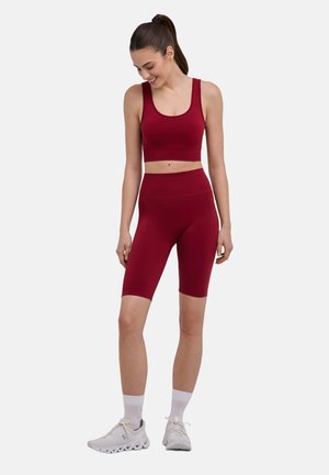 Junge Frau in rotem Sport-BH und hoch taillierten Radlerhosen, weißen Socken und Turnschuhen, steht und schaut lächelnd nach unten.