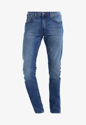 Wąskie niebieskie dżinsy o średnim stanie, pięć kieszeni i zapięcie na brązowy guzik. Materiał ma lekko wyblakłe wykończenie.