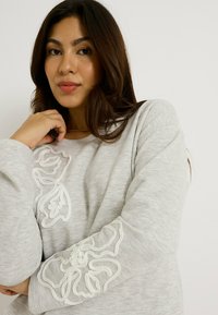 Sudadera gris claro con diseños florales bordados en blanco en la parte delantera y las mangas; textura suave y corte casual.