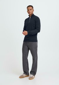 Pull à zip bleu marine avec des manches longues, tissu texturé ; associé à un jean droit gris foncé et des baskets grises avec des rayures blanches.