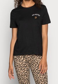 Vrouw in een zwart T-shirt met de tekst "GA LOS!" en een eikelafbeelding, gecombineerd met beige en zwarte panterprint broek.