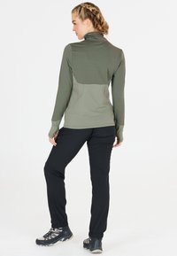 Langärmliges, olivgrünes Oberteil mit hohem Kragen und strukturiertem Rücken; kombiniert mit schwarzen, schmal geschnittenen Hosen. Verstärkte Nähte und elastische Bündchen.