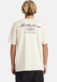Een persoon met blond haar die een lichtbeige Billabong t-shirt draagt met een kruisende surfplank graphic en de tekst "Boardshorts USA AUS EUR JPN" op de rug.