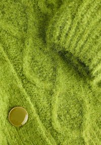 Tessuto di maglia verde brillante con una texture pelosa. Presenta un polso a coste e un bottone rotondo verde come accentuazione. Regolabile e con un intreccio a più fantasie.
