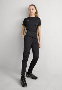 Fekete sportos ruházat arany díszítéssel; rövid ujjú felső és szűk fazonú nadrág sima anyagból. Fekete focicipő fehér részletekkel.