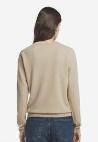 Beżowy sweter z długimi rękawami, okrągłym dekoltem, ribbowymi mankietami i dołem, wykonany z miękkiego materiału, noszony z niebieskimi dżinsami.