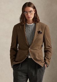 Junge Person mit schulterlangem Haar und runden Brillen trägt einen braunen Blazer über einem grauen Zopfpullover und dunkle Hose, Hände in den Taschen.