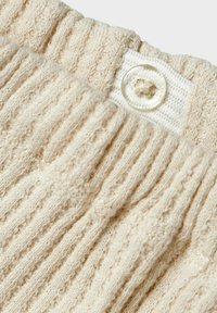 Beige ribbet strikket stof med en tekstureret overflade, der har et hvidt vævet mærke og en klar knapdetalje på taljen.