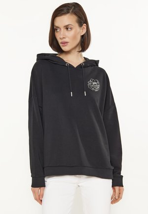 Vrouw draagt een zwarte hoodie met een klein wit bloemmotief op de borst, gecombineerd met een witte broek, kijkt opzij.