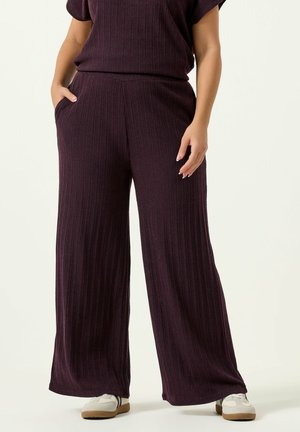 Frau trägt dunkelviolette, gerippte, weit geschnittene Hosen und weiße Turnschuhe, steht mit einer Hand in der Tasche vor einem schlichten Hintergrund.