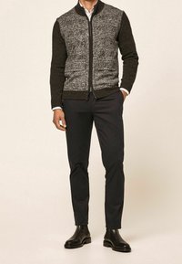 Uomo che indossa un maglione scuro con zip e una parte frontale grigia a fantasia, pantaloni sottili neri, stivali neri e una camicia bianca sotto.