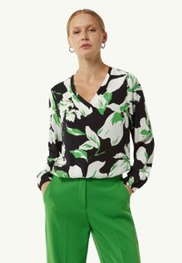 Blumenbluse in Schwarz, Weiß und Grün mit langen Ärmeln, V-Ausschnitt und gerafften Manschetten. Kombiniert mit grünen Hosen.