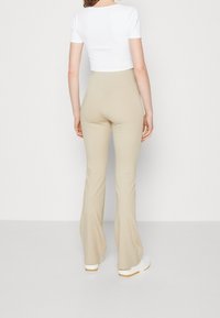 Žena oblečená v béžových rozšířených kalhotách, bílém krátkém topu a bílých teniskách, stojící zády k jednobarevnému bílému pozadí.