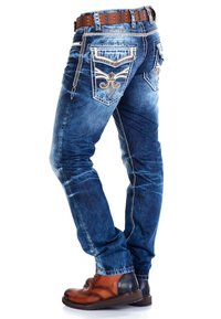 Ausgeblichene blaue Jeans mit bestickten Gesäßtaschen, die ein florales Design zeigen. Kombiniert mit braunen Schnürstiefeln und einem Nieten-Gürtel.
