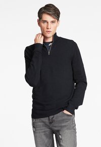 Dunkelblauer Zip-Kragen-Pullover aus Strickstoff, ausgestattet mit gerippten Bündchen und Saum, getragen zu grauen, used-look Jeans. Glatte Textur und figurbetonter Schnitt.