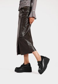 Scarpe platform in vernice nera con suola spessa e dettaglio a fibbia rettangolare, abbinate a una gonna di pelle lucida marrone fino al ginocchio.