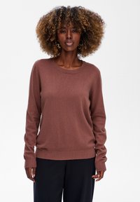 Brązowy, długi sweter z miękkiej dzianiny, z okrągłym dekoltem i prą�żkowanymi mankietami, w zestawie z ciemnymi spodniami. Minimalistyczny design, bez wzorów.