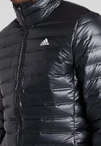 Sort puffjakke med vandret quiltning, høj krave og lynlås. Har hvidt Adidas-logo på venstre bryst. Glat, skinnende materiale.