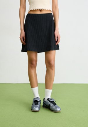 Žena v černé žebrované mini sukni, bílých žebrovaných ponožkách, šedých teniskách Nike a krátkém bílém topu stojí na zelené podlaze.