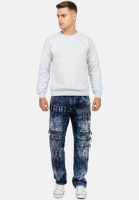 Felpa grigia con collo a giro, abbinata a jeans cargo blu strappati con più tasche, cerniere e motivi. Sneakers bianche.