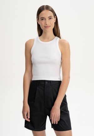 Jeune femme aux longs cheveux bruns portant un crop top blanc sans manches et un short noir taille haute, debout devant un fond uni.