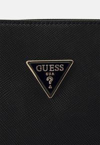 Sort tekstureret materiale med en sort trekantet Guess USA-logo plade med guldkant, centreret på overfladen.