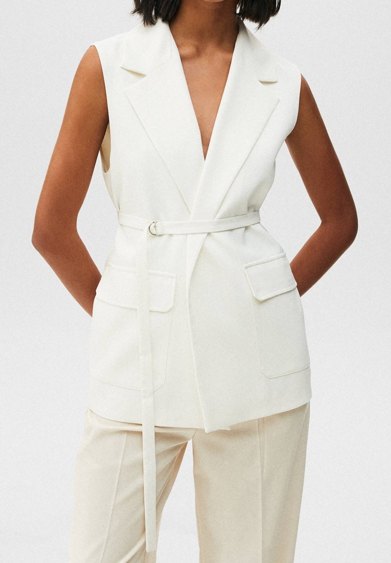 Ärmelloser weißer Blazer mit tiefem V-Ausschnitt, Taillengürtel und zwei Fronttaschen; gefertigt aus strukturiertem Stoff mit glatter Textur.