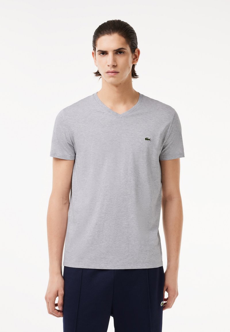 Szary t-shirt z krótkim rękawem w kształcie litery V, wykonany z miękkiego materiału, z małym zielonym logo krokodyla na lewej piersi. Prosty i klasyczny design.