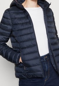 Donna con giacca trapuntata blu scuro con cappuccio sopra una camicia bianca e jeans blu scuro, con una mano nella tasca della giacca.