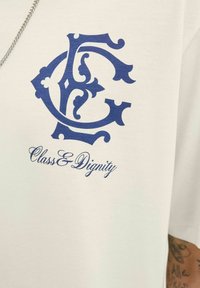 Bílá košile s ozdobným modrým monogramem "C" a "D" nad kurzívními nápisy "Třída a Důstojnost," kterou nosí osoba s viditelným tetováním na paži.