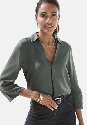 Blusa verde scuro con colletto a bottoni e maniche a tre quarti, abbinata a una gonna nera e accessori dorati, comprese collane a strati.