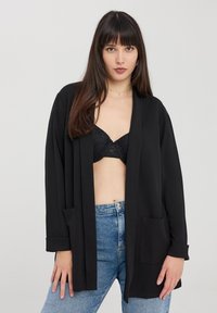 Cardigan lungo nero realizzato in tessuto leggero, con apertura davanti, due tasche anteriori e risvolti, indossato sopra un reggiseno nero e jeans.