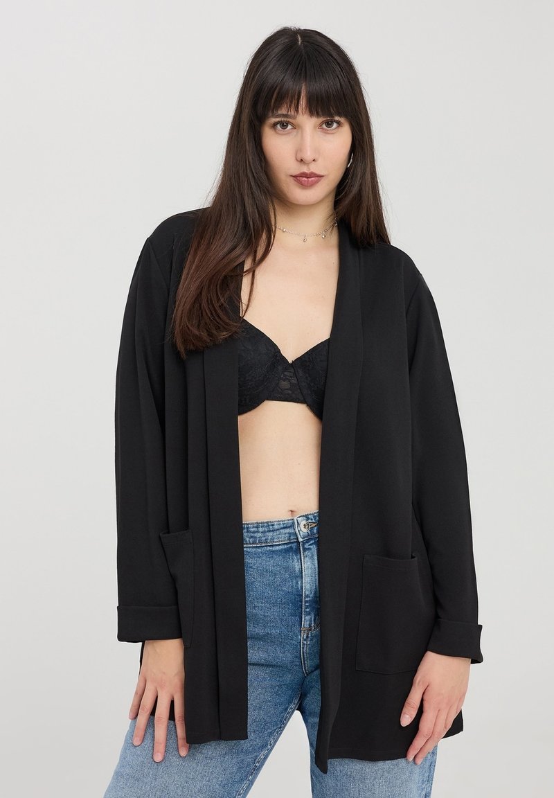 Cardigan lungo nero realizzato in tessuto leggero, con apertura davanti, due tasche anteriori e risvolti, indossato sopra un reggiseno nero e jeans.