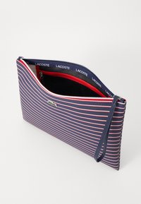 Clutch torba u mornarsko plavoj i crvenoj prugastoj boji, napravljena od teksturirane tkanine. Ima zatvaranje na munje, unutarnju džep, i narukvicu s Lacoste logotipom.