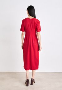 Sarkans midi kleita ar īsām piedurknēm, struktūrētu augšdaļu un slēgtu rāvējslēdzēju aizmugurē. Modelis apvieno to ar tumši brūnām augstpapēžu kurpēm.