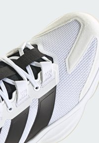 Baskets de sport blanches avec une tige en mesh respirant, des accents noirs, des panneaux texturés et des lacets plats blancs sur une sangle centrale noire.