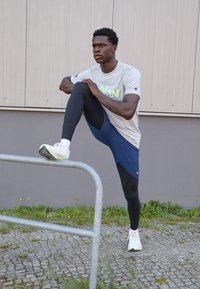 Conjunto deportivo que incluye una camiseta gris de manga corta con texto verde neón, pantalones cortos azul marino, leggings negros y zapatillas blancas, situado junto a una barandilla.