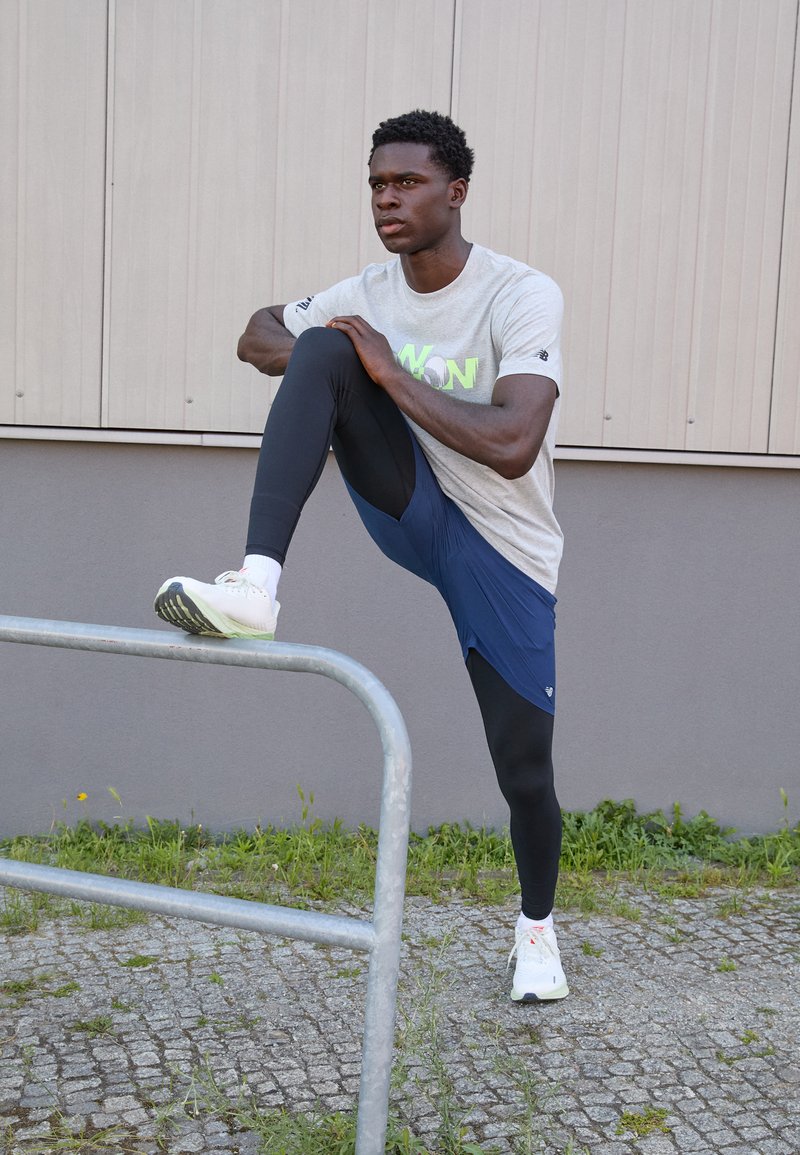 Tenue de sport comprenant un t-shirt gris à manches courtes avec un texte vert fluo, un short bleu marine, des leggings noirs et des baskets blanches, positionnée près d'une rambarde.